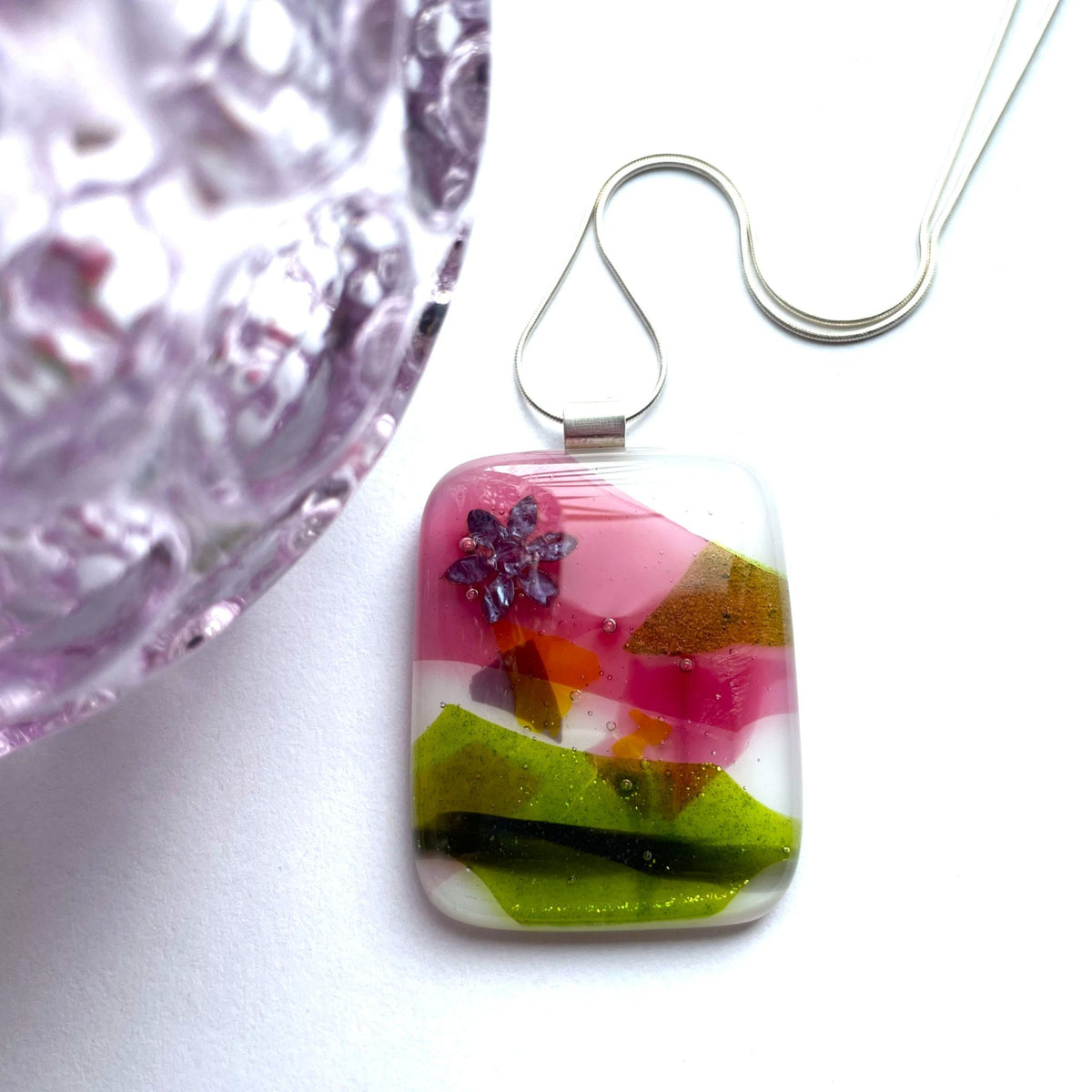Fused glass pendant with pink, purple and green confetti glass displayed against colourful glass