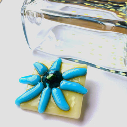 Front view of a warm vanilla fused glass brooch with a cool blue flower and teal green centre